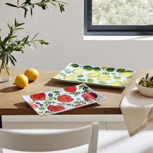 Set di vassoietti in ceramica artigianale decorati: uno con limoni e foglie verdi, l’altro con pomodori rossi e olive su sfondo bianco. Disposti su un tavolo in legno in un ambiente luminoso e minimalista, con luce naturale da finestra, accompagnati da limoni freschi, una pianta di ulivo e una ciotola di olive.