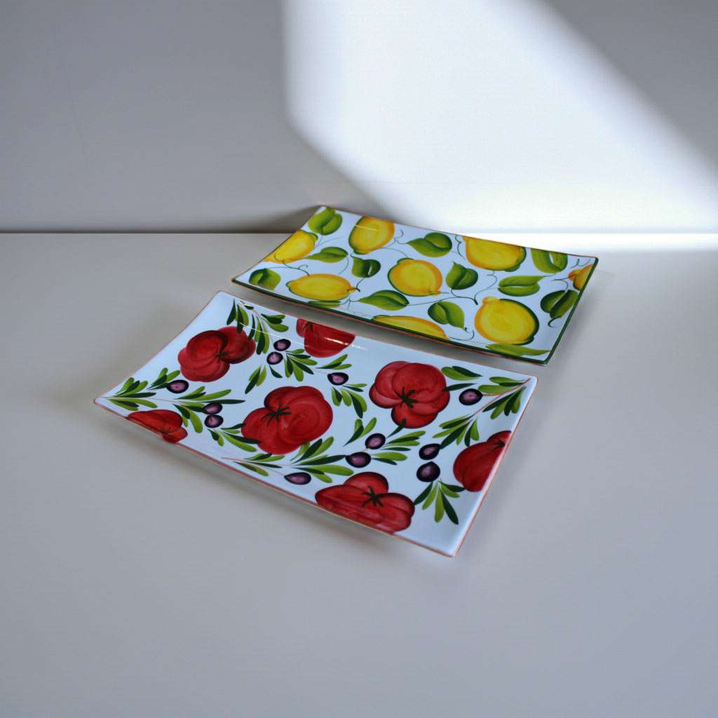 Set di vassoietti in ceramica artigianale decorati a mano: uno con limoni e foglie verdi, l’altro con pomodori rossi e olive su sfondo bianco. Disposti su una superficie chiara in un ambiente minimalista, illuminati da luce naturale con ombre morbide.